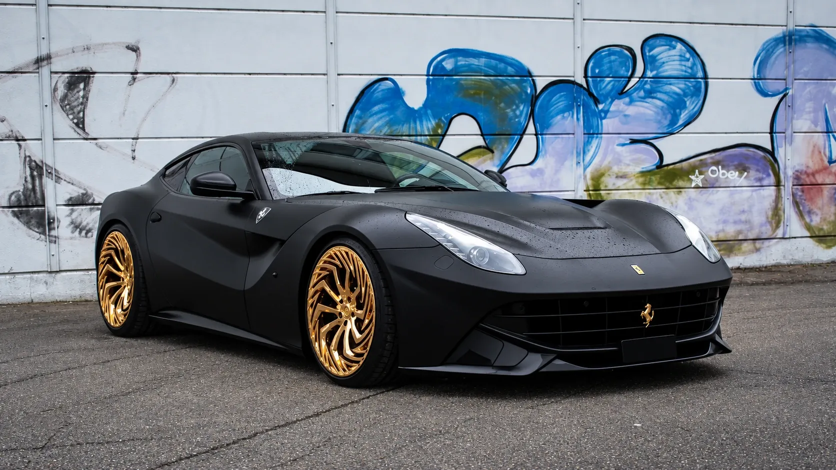 Matte black Ferrari on gold directional-spoke wheels in front of a painted wall